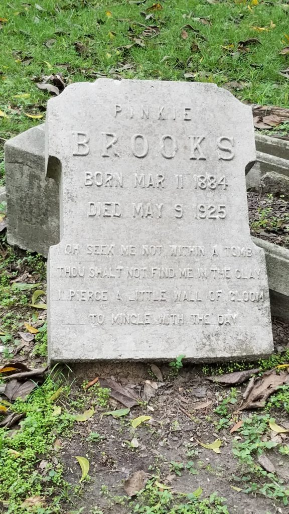 Gravestone propped upon another stone with grass and leaves below. Reads: "Pinkie Brooks; Born Mar 11, 1884; Died May 9, 1925. The rest of the text is difficult to read.