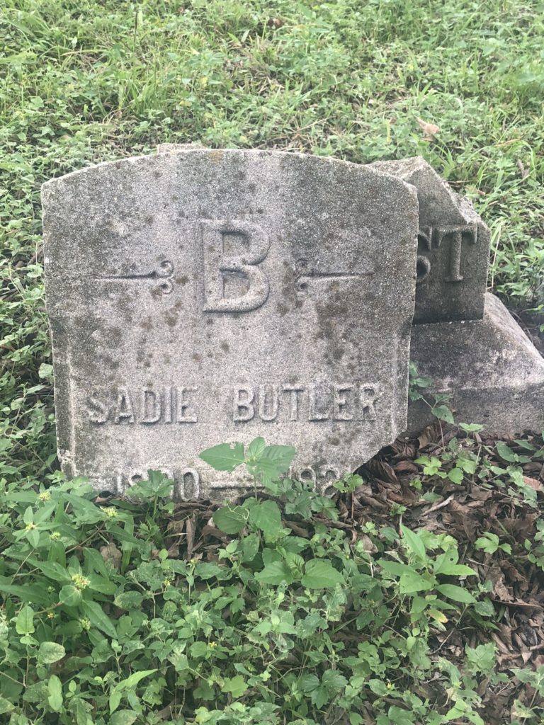 Headstone is in pieces. The piece in front has a big "B" at the top and reads "Sadie Butler." The years, written below are not legible because they're broken off from the bottom of the headstone which sits behind this piece.